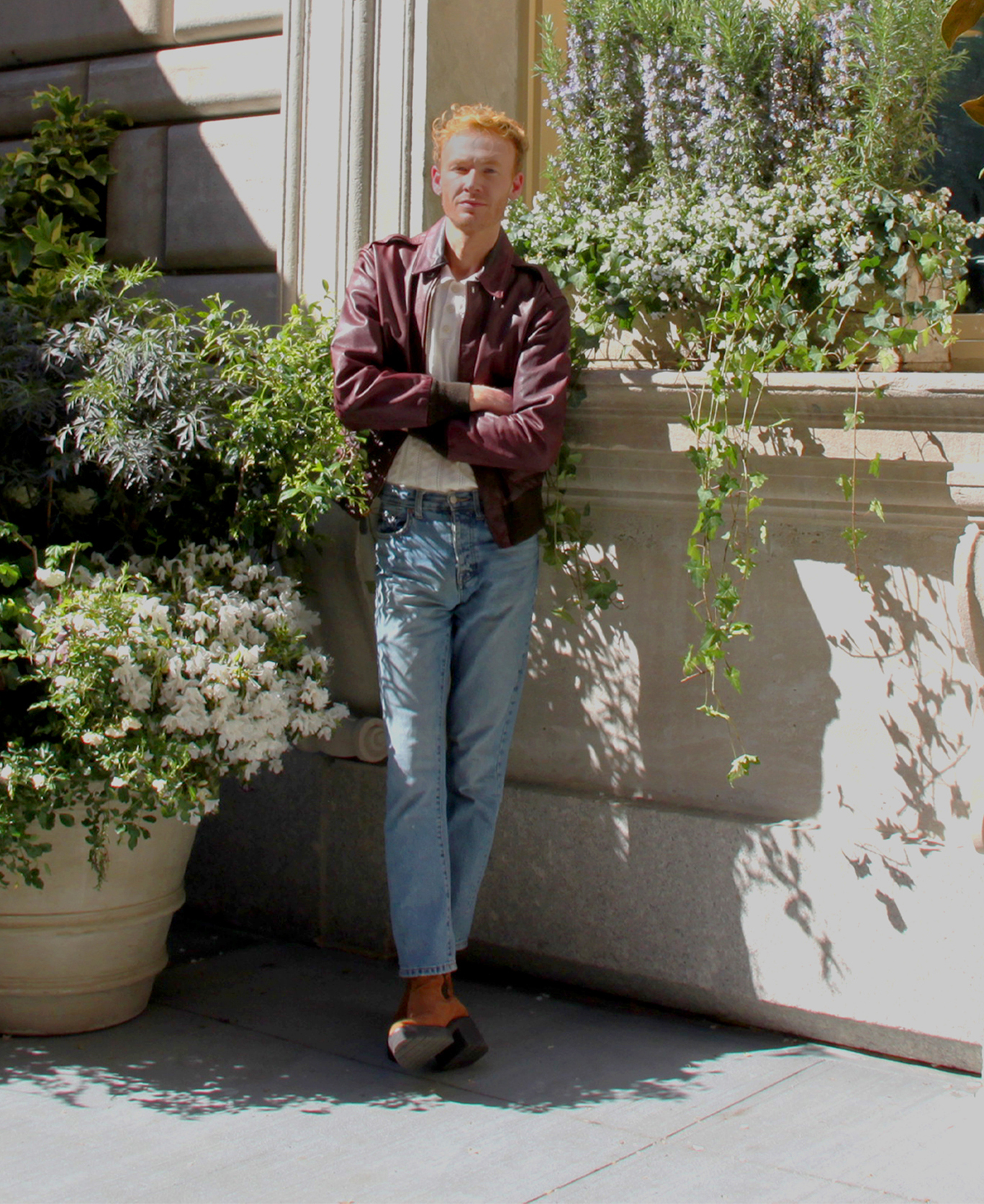 A man in a brown jacket poses beside a large flower pot for a photo.