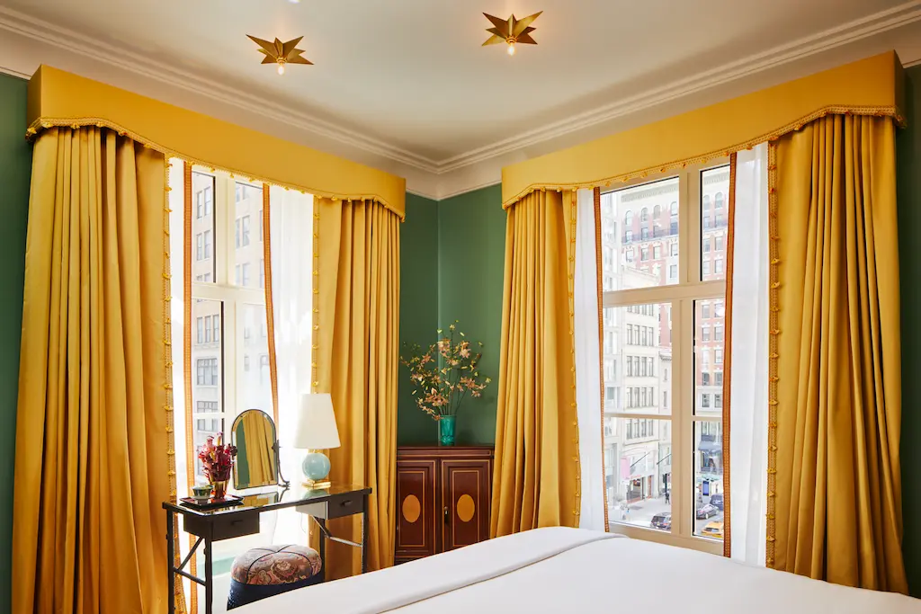 A well-organized room with a cozy bed and a table by the window, covered with a yellow curtain.