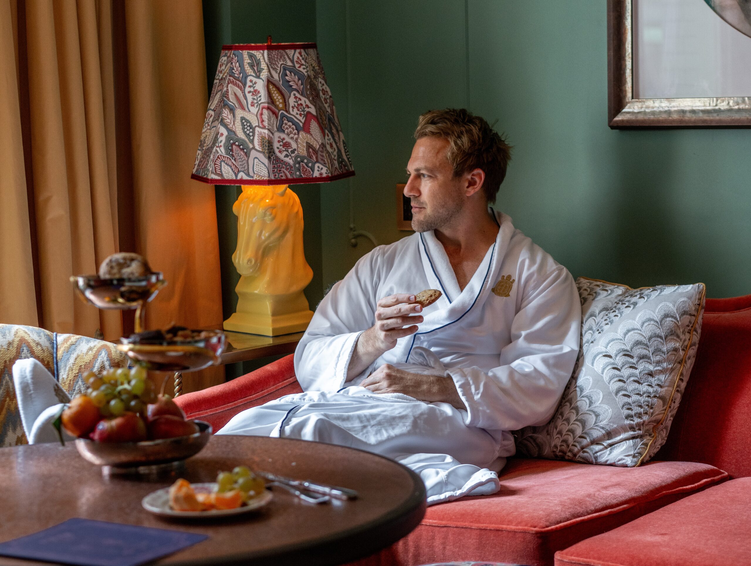 A man in a bathrobe relaxes on a cozy sofa, enjoying breakfast while looking away.