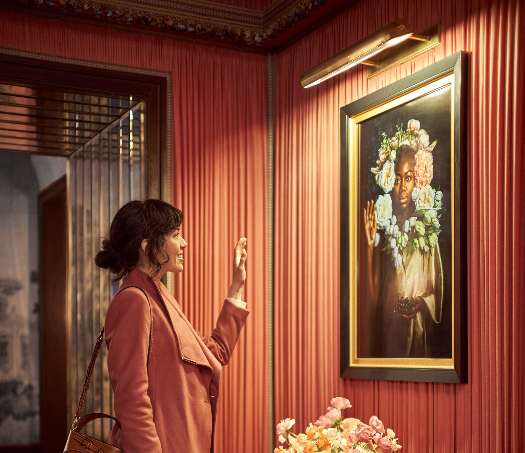 A lady in a pink jacket raises her hand toward the beautiful wall-mounted portrait.