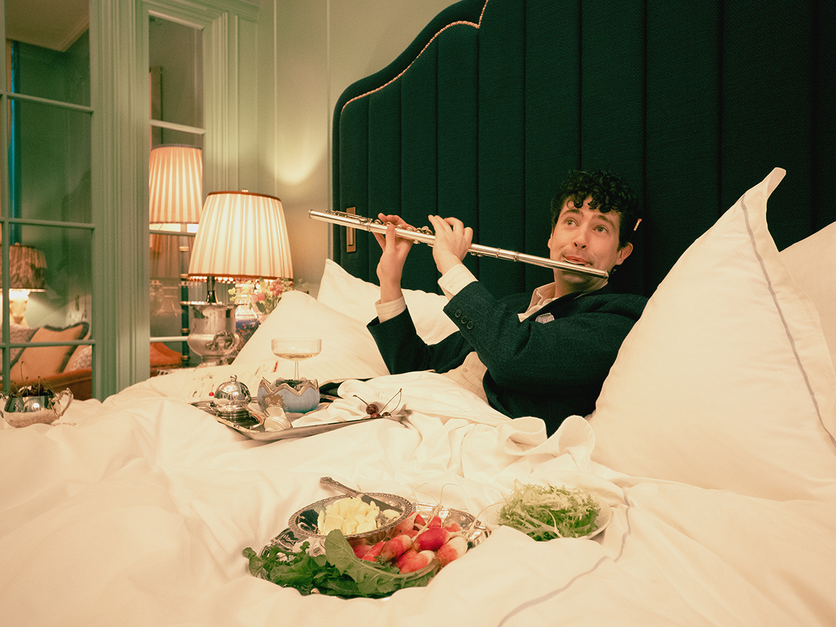 A well-suited man playing the flute in bed with a breakfast tray nearby.