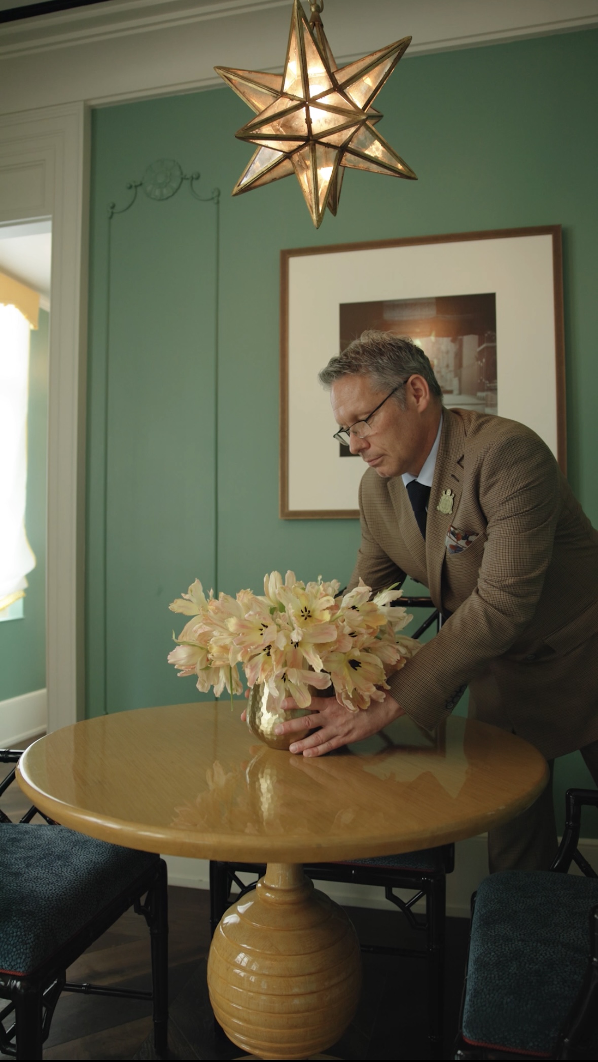 A man in a brown suit arranges a vase of light pink flowers on a small wooden table.